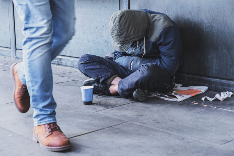 Homeless man sitting on the street in the shadow of the building and begging for help and money. Problems of big modern cities. Image symbolizing addiction and homelessness